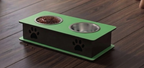 A pet elevated feeder with two bowls, one containing dry food, placed on a dark wood floor. The feeder has a green top and base with a black side panel featuring black paw print cutouts, and is paired with a stainless steel bowl.