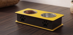 A two-bowl pet feeder and waterer stand with a yellow top tray, black base, and stainless steel bowls, shown on a dark wooden table.