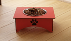 A red elevated dog feeder with a metal bowl filled with kibble, placed on a light wood floor. The feeder has a black paw print cutout on the front.