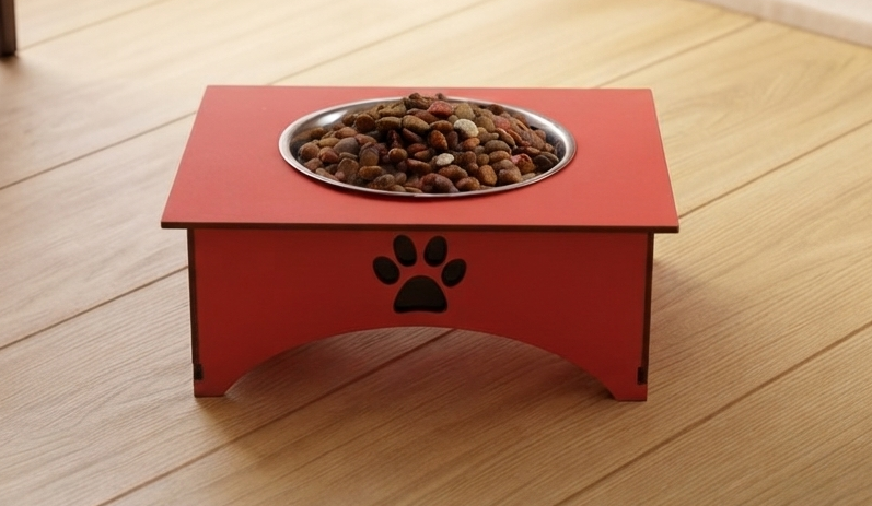 A red elevated dog feeder with a metal bowl filled with kibble, placed on a light wood floor. The feeder has a black paw print cutout on the front.