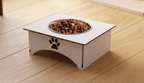 A white elevated pet feeder with a stainless steel bowl filled with dry kibble, placed on a light wood floor. The feeder has a black paw print cutout on the front.