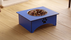 A blue elevated pet food bowl stand with a metal bowl filled with kibble, placed on a light wood floor. The stand has a black paw print cutout on the front.