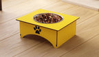 Yellow elevated pet feeder with a stainless steel bowl filled with dry kibble, placed on a light wood floor. The feeder has a black paw print graphic on the front.