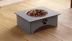 A square elevated pet feeder with a stainless steel bowl filled with kibble, placed on a light wood plank floor. The feeder is a muted gray color with a black paw print cutout on the front panel.