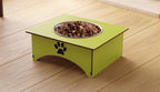 A lime green elevated square dog feeder with a stainless steel bowl filled with kibble, placed on a light wood floor. The feeder features a black paw print graphic on the front side.