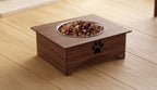 A square elevated wooden pet feeder with a stainless steel bowl filled with kibble, placed on a light wood floor. The feeder has a black paw print cutout on the front.