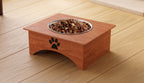 A wooden elevated pet feeder with a stainless steel bowl filled with dog kibble, placed on a light wood plank floor. The feeder has a black paw print engraving on the front.
