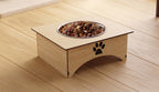 A wooden elevated pet feeder with a stainless steel bowl filled with kibble, placed on a light wood floor. The feeder has a black paw print logo on the front.