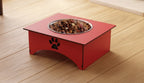 A red elevated pet food bowl with a stainless steel insert filled with dry dog food, placed on a light wood floor. The bowl features a black paw print cutout on the front side.