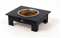 A black elevated pet feeder stand with a stainless steel bowl filled with dry kibble, photographed on a clean white background.