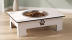 A white elevated pet feeder table with a stainless steel bowl filled with dog food, set on a light wood floor against a pale green wall.