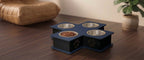 A four-bowl elevated pet feeder and waterer with a dark blue cross-shaped base, stainless steel bowls, and gold paw print details, shown on a wooden table in a home setting.
