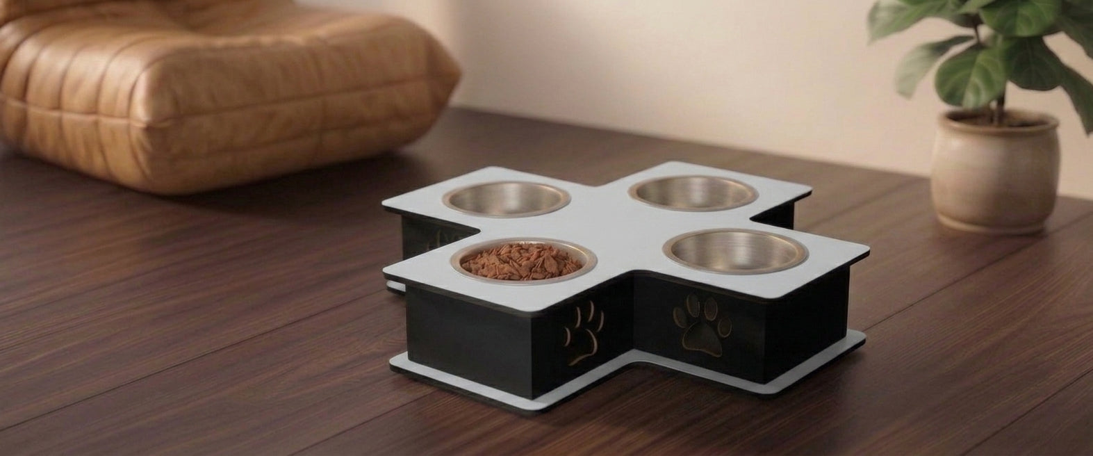 A four-bowl elevated pet feeder and waterer with a black and white cross-shaped base, shown on a dark wooden table in a home setting with a cushion and potted plant in the background.