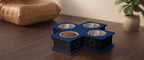 A four-bowl elevated pet feeder and waterer with a blue top and black base, featuring a paw print design on the front, set on a dark wood floor.