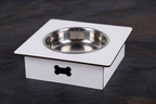A white square pet feeder or elevated bowl stand with a stainless steel bowl set inside, featuring a black bone-shaped cutout on the front, photographed on a dark textured surface.