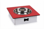 A square pet feeder or elevated bowl stand with a red top surface, a stainless steel bowl, and a white base featuring a black bone-shaped cutout, shown on a white background.