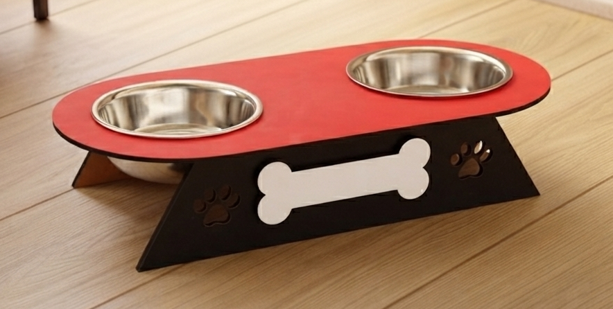 A two-bowl elevated pet feeder and water bowl stand with a red top platform, stainless steel bowls, a black angled base, a white bone-shaped center detail, and paw print cutouts, shown on a light wood floor.