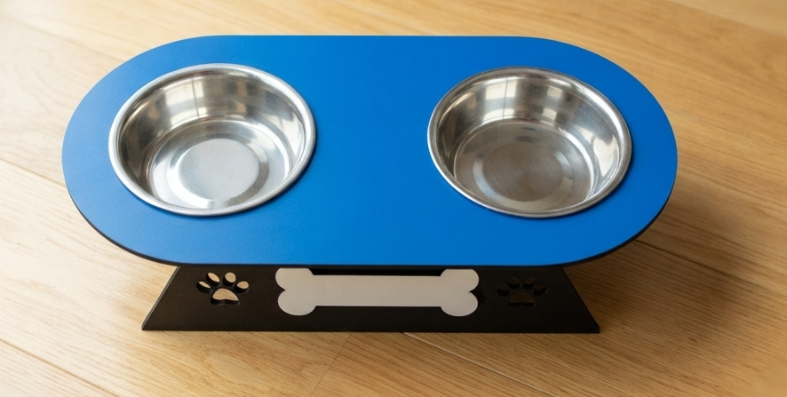 A blue elevated pet feeder and water bowl stand with two stainless steel bowls, shown on a light wood floor. The stand has a black base with bone and paw print cutouts.