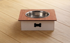 A pet elevated feeder with a square wooden top, a stainless steel bowl, and a white base with a black bone-shaped cutout, shown on a light wood plank floor.