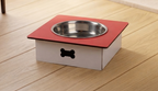 A raised pet feeder and water bowl stand with a red top platform, a stainless steel bowl, and a white base featuring a black bone-shaped handle, shown on a light wood floor.