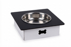 A square pet feeder or elevated bowl stand with a black top surface, a stainless steel bowl, and a white base with a black bone-shaped cutout, shown on a white background.