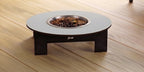A low-profile pet fire table with a round light gray top, dark brown wooden legs, and a stainless steel fire bowl filled with wood chips, set on a light wood plank floor.