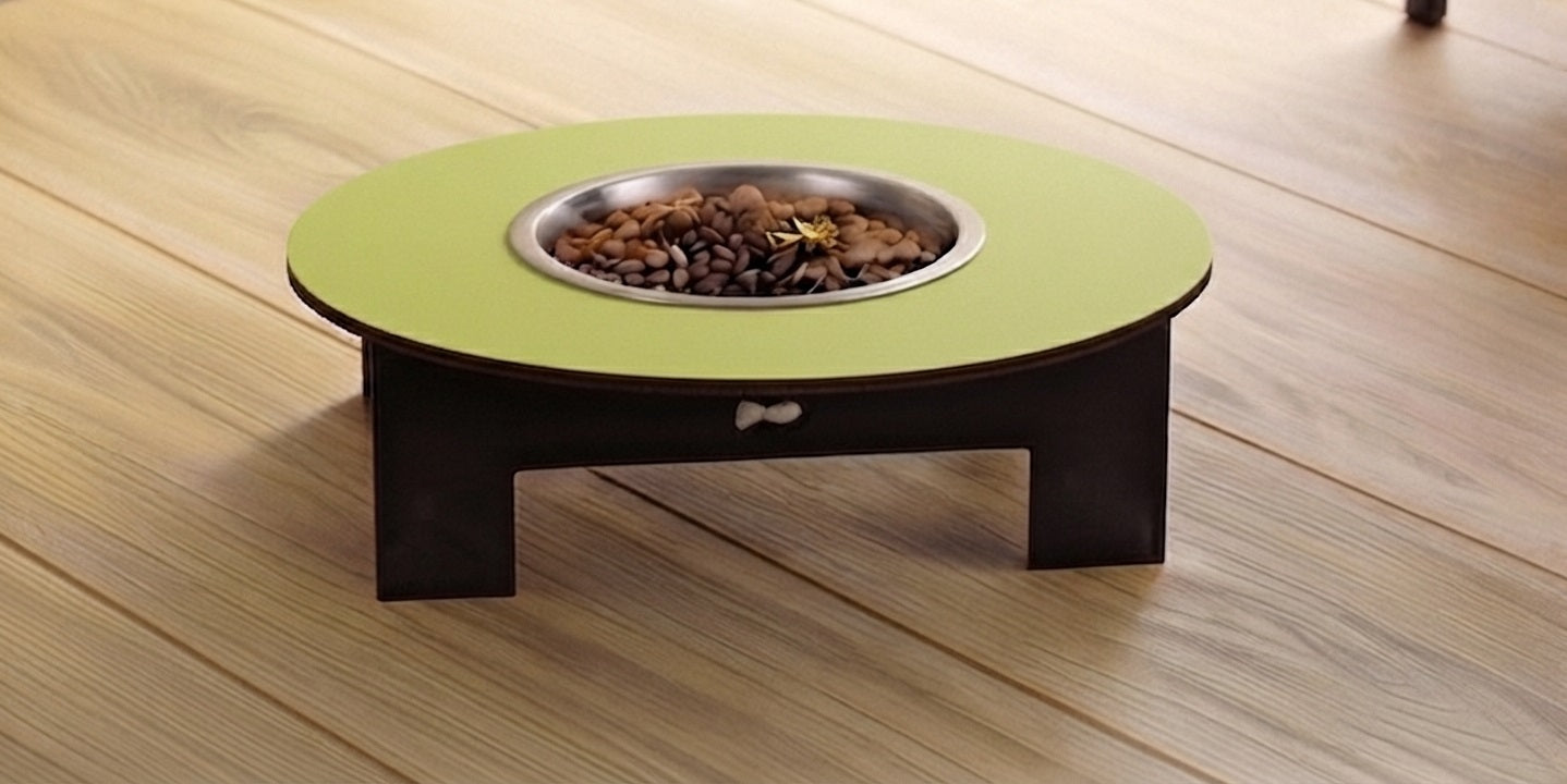 A round elevated pet fire table with a light green top, dark brown wooden legs, and a stainless steel bowl filled with dog food, set on a light wood plank floor.