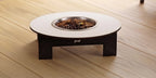 A low-profile elevated pet feeder table with a round white top, dark brown wooden legs, and a stainless steel bowl filled with kibble, set on a light wood plank floor.