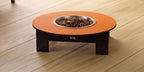 A low-profile round pet fire pit table with a terracotta orange top, dark brown wooden legs, and a stainless steel bowl filled with burning coals, set on a light wood plank floor.