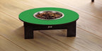 A raised pet feeder table with a bright green circular top, dark brown wooden legs, and a stainless steel bowl filled with kibble, set on a light wood plank floor.