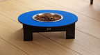 A raised pet feeder table with a bright blue circular top, dark wooden legs, and a stainless steel bowl filled with kibble, placed on a light wood plank floor.