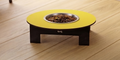 A raised pet feeder table with a bright yellow circular top, a stainless steel bowl filled with kibble, and a dark wooden base, shown on a light wood plank floor.