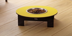 A raised pet feeder table with a bright yellow circular top, a stainless steel bowl filled with kibble, and a dark wooden base, shown on a light wood plank floor.