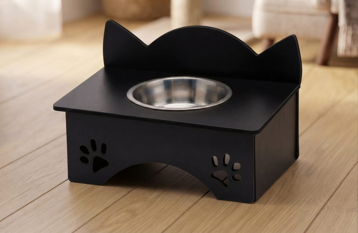 A black cat pet feeder stand with a stainless steel bowl, set on a light wood floor in a softly blurred home environment.