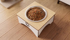 A raised wooden pet feeder stand with a stainless steel bowl filled with dry kibble, placed on a light wood floor.