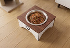 An elevated pet feeder with a square wooden top, white base, and a stainless steel bowl filled with dry cat food, placed on a light wood floor.