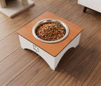 A square elevated pet feeder with a light brown wood-grain top and white base, featuring a stainless steel bowl filled with dry cat food, placed on a dark wood floor.