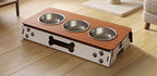 A pet elevated feeding station with a wood-grain top, three stainless steel bowls, and a white base decorated with black paw prints and a bone-shaped handle, shown on a light wood floor.