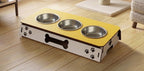 A pet elevated feeding station with three stainless steel bowls on a yellow and white wooden base, shown on a light wood floor in a home setting.