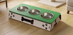 A three-bowl elevated pet feeder and water fountain with a green top surface, stainless steel bowls, and a white base decorated with black paw prints and a bone cutout, shown on a wooden floor.