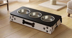A pet elevated feeding station with three stainless steel bowls on a black top, set on a white base decorated with paw prints and a bone-shaped cutout, shown on a wooden floor.