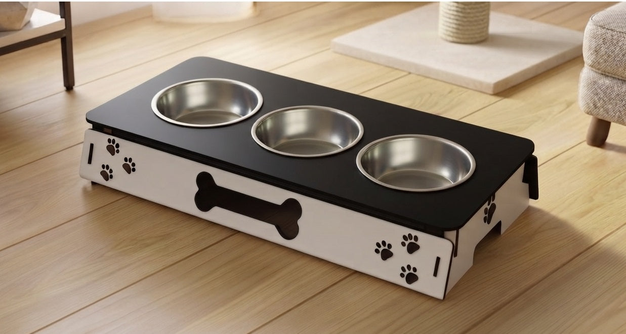 A pet elevated feeding station with three stainless steel bowls on a black top, set on a white base decorated with paw prints and a bone-shaped cutout, shown on a wooden floor.