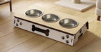A wooden elevated pet feeder and water bowl stand with three stainless steel bowls, shown on a light wood floor in a home setting.