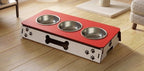 A raised pet feeder and water bowl stand with three stainless steel bowls on a red top, set on a light wood floor in a home environment.