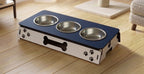 A pet elevated feeding station with three stainless steel bowls, featuring a navy blue top and a white side panel decorated with black paw prints and a bone silhouette, shown on a wooden floor.