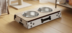 A wooden elevated pet feeder and water bowl stand with two stainless steel bowls, shown on a light wood floor in a home setting.