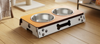A pet elevated feeding station with two stainless steel bowls, a light wood top, and a white base decorated with black paw prints and a bone graphic, shown on a wooden floor.