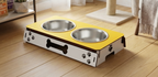 A pet elevated feeding station with two stainless steel bowls, featuring a yellow top surface, white side panels with black paw prints and a bone-shaped handle, placed on a wooden floor in a home setting.