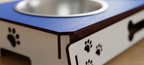 Close-up of a pet elevated feeder or bed with a blue top, white sides, dark brown trim, a stainless steel bowl, and black paw print cutouts.