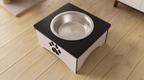A square elevated pet feeder or dish stand with a black top, white sides, a stainless steel bowl, and a paw print cutout on the front, shown on a light wood floor.